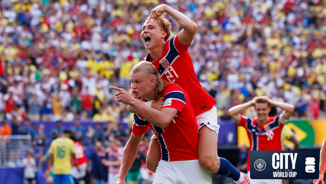 Two football players in red Norway jerseys celebrate a goal, with one player piggybacking on the other. A crowd and other players in similar jerseys are blurred in the background. City World Cup logo is visible.
