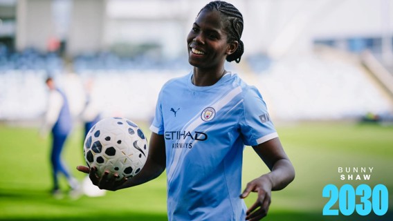 A person wearing a Manchester City jersey holding a soccer ball on a field. The jersey features the Manchester City crest and sponsors.