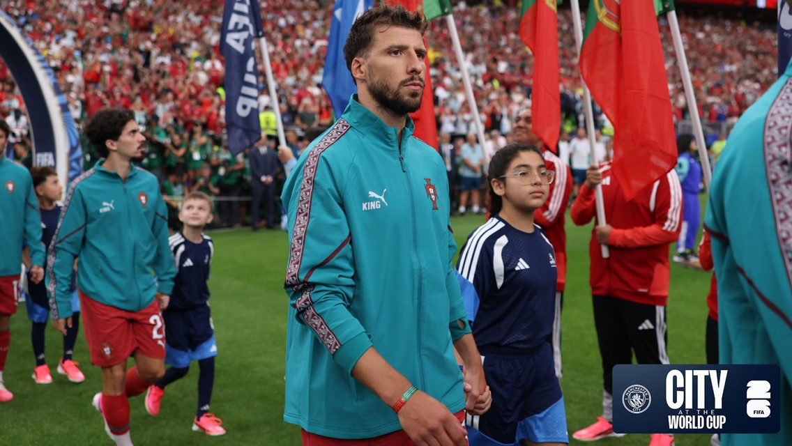 Football players wearing tracksuits with national flags in a stadium setting, accompanied by children with similar attire. Crowd visible in the background, with a City World Cup logo.