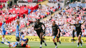 Soccer players in action during a match, one team in black and another in blue and white, with a goalkeeper on the ground and a crowd in the stadium.