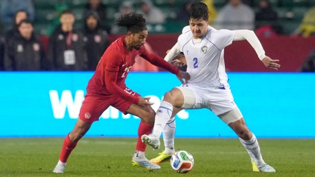 Players from two teams are engaged in a soccer match, battling for the ball on the pitch. One player is in red and the other in white, with a focused attempt to control the ball.