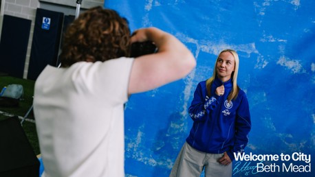 A person is posing in front of a blue backdrop during a photoshoot. They are wearing a blue jacket with a badge on it. The backdrop includes the text 'Welcome to City Beth Mead'.
