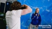 A person is posing in front of a blue backdrop during a photoshoot. They are wearing a blue jacket with a badge on it. The backdrop includes the text 'Welcome to City Beth Mead'.