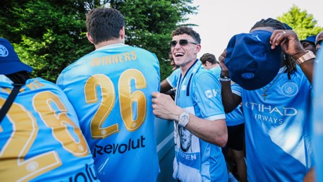 Manchester City fans celebrating, wearing blue jerseys, with some holding caps. One jersey has 'Winners 26' written on it.