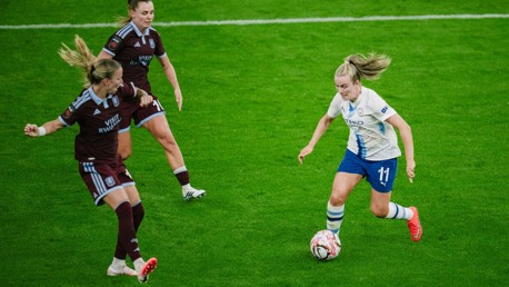 Action shot of three female soccer players on the field during a match. One in white attempts to maneuver the ball with two in maroon jerseys nearby.