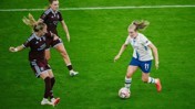 Action shot of three female soccer players on the field during a match. One in white attempts to maneuver the ball with two in maroon jerseys nearby.