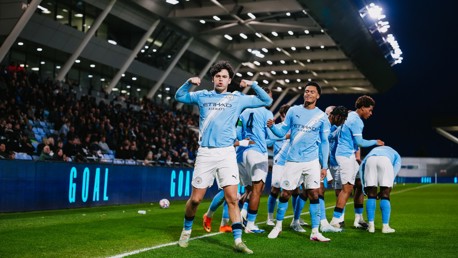 Soccer players in Manchester City jerseys celebrating a goal on the field with a crowd in the background.