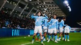 Soccer players in Manchester City jerseys celebrating a goal on the field with a crowd in the background.