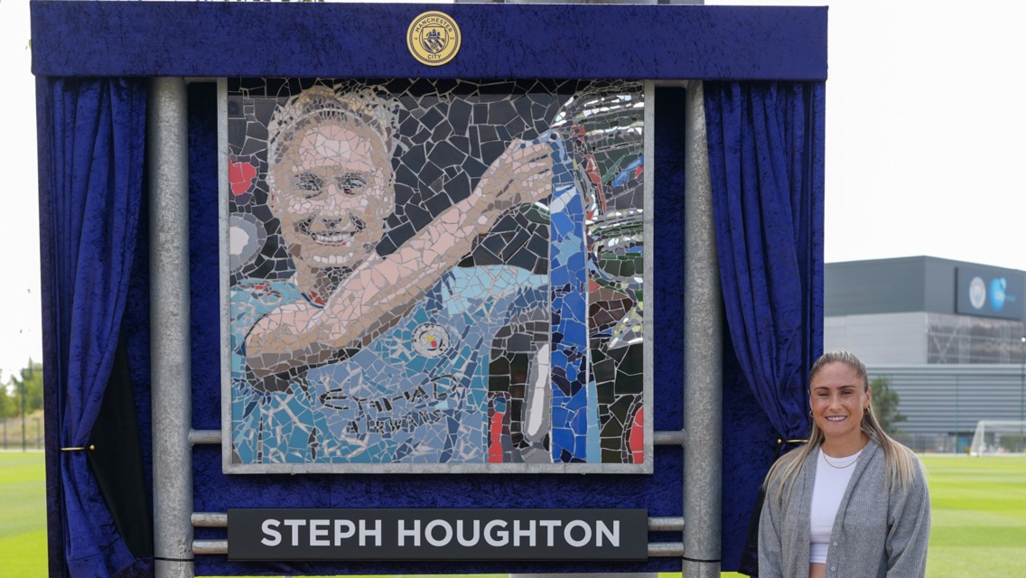 A person stands beside a mosaic artwork depicting someone in a Manchester City jersey holding a trophy, with a name plaque reading 'Steph Houghton.'