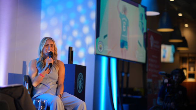 A member of the Manchester City Women's team is giving an interview on stage with a microphone. Behind her, there is a large screen displaying a Manchester City player waving.