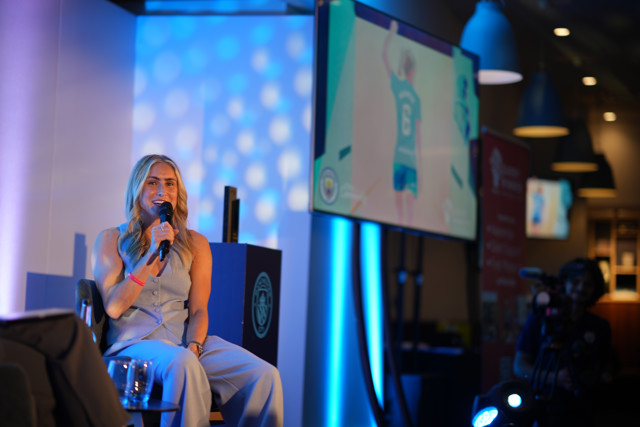 A member of the Manchester City Women's team is giving an interview on stage with a microphone. Behind her, there is a large screen displaying a Manchester City player waving.