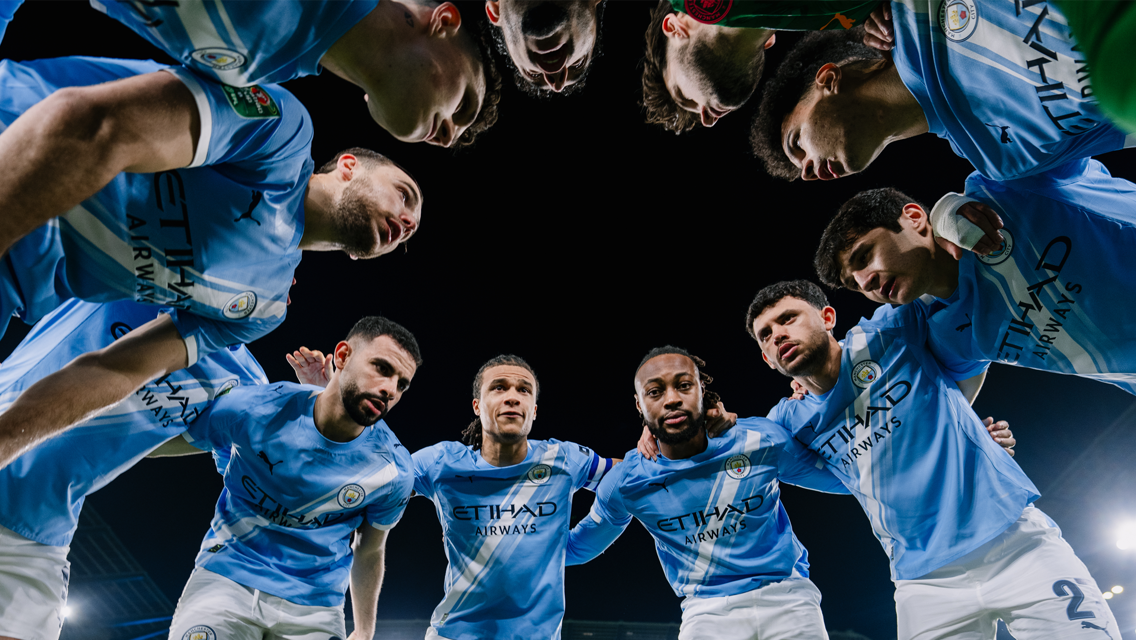 Manchester City players in a huddle, wearing light blue kits with Etihad Airways branding, viewed from a low angle at night.