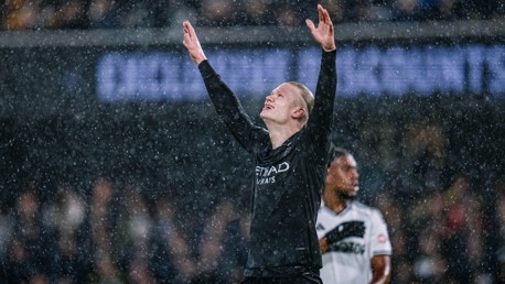 A Manchester City player celebrating in the rain with arms raised, wearing a black kit with visible 'Etihad Airways' logo.