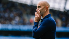 A man in a blue jacket stands in a side profile view, with his fingers touching his lips in a thoughtful pose.