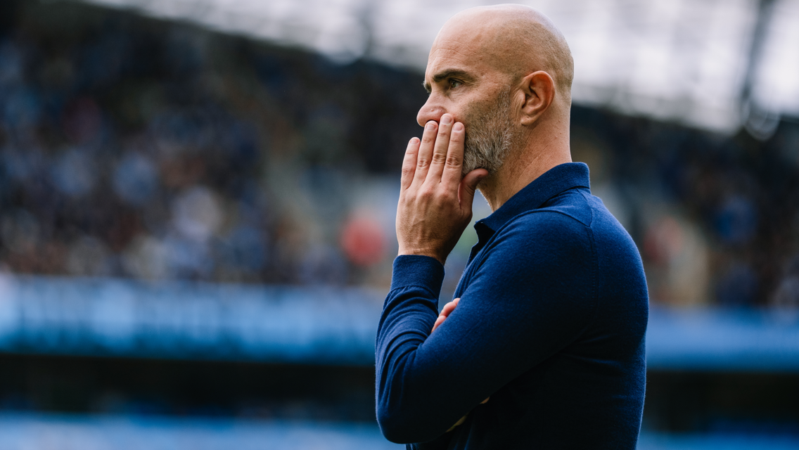 A man in a blue jacket stands in a side profile view, with his fingers touching his lips in a thoughtful pose.
