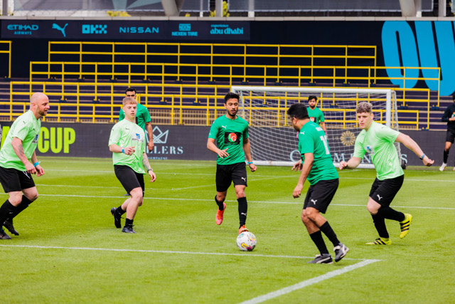 A group of amateur players in green and light green kits engaging in a soccer match on a playing field.