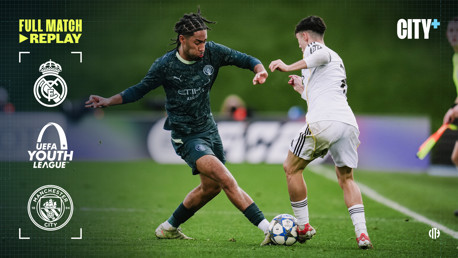 Real Madrid and Manchester City youth teams compete in the UEFA Youth League with a Manchester City player challenging a Real Madrid player for the ball.