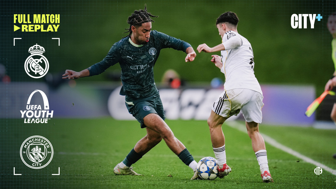 Real Madrid and Manchester City youth teams compete in the UEFA Youth League with a Manchester City player challenging a Real Madrid player for the ball.