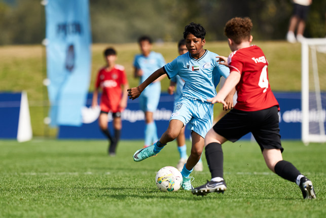 Youth soccer players in action during a competitive match, with one player in a blue uniform dribbling the ball while being challenged by another player in a red uniform.