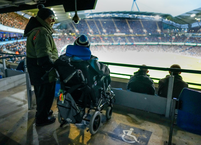 Two people, one in a wheelchair and another standing, watching a match at a stadium from an accessible viewing area.