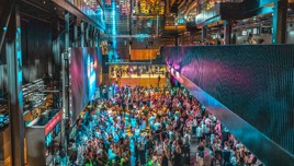 A large crowd gathered inside a modern venue with colorful lighting, a big screen, and multiple levels.