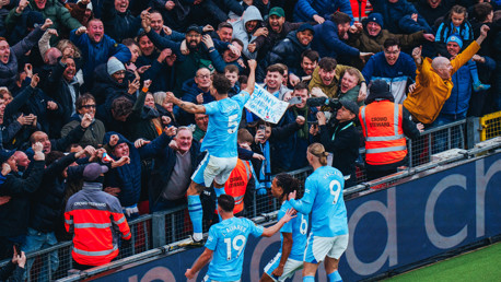 Manchester City players celebrating a goal with fans; a player holding a sign, surrounded by excited spectators and stewards.