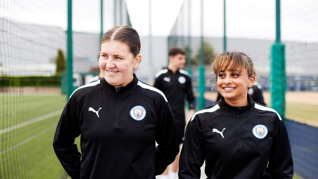Two Manchester City Women's players in training gear with club crest, walking on a football pitch at training ground.