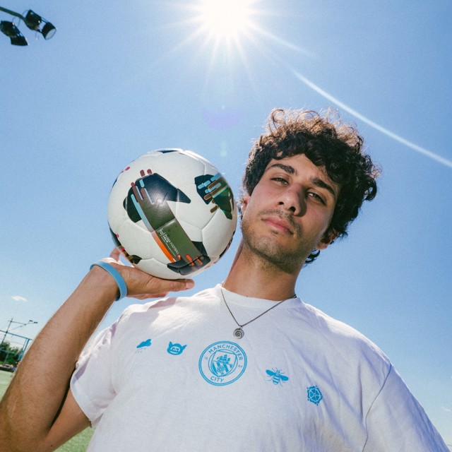 A young man holding a soccer ball with a Manchester City logo shirt under a bright sun.