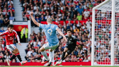 A football player in a red jersey defends against a light blue jersey player attempting to score a goal, with a goalkeeper poised to react, in front of a crowded stadium.
