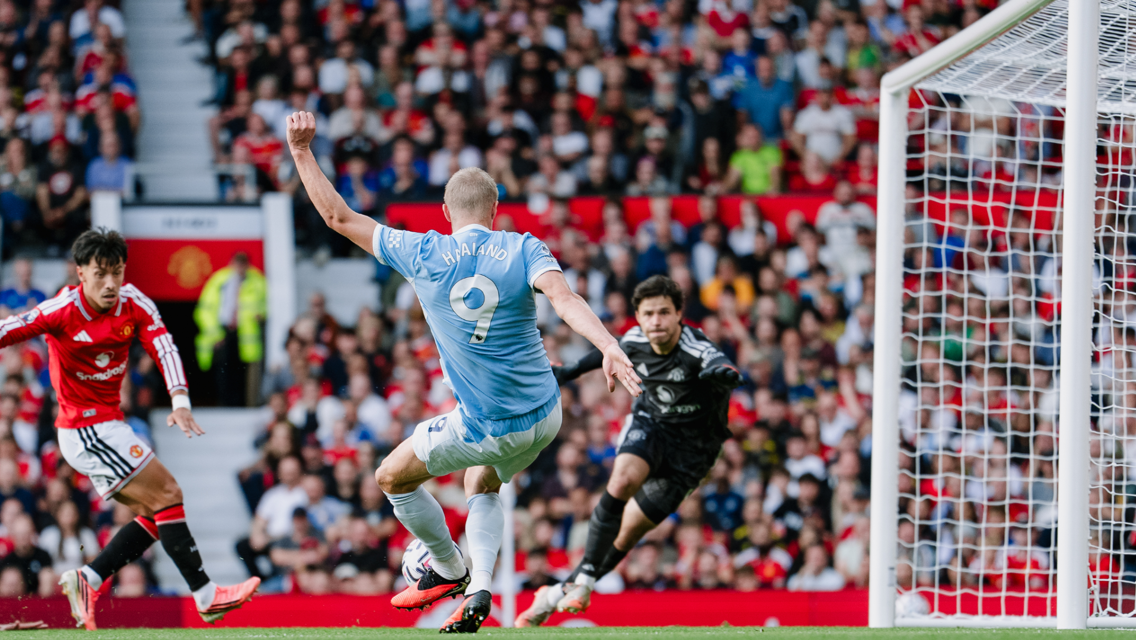 A football player in a red jersey defends against a light blue jersey player attempting to score a goal, with a goalkeeper poised to react, in front of a crowded stadium.