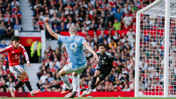 A football player in a red jersey defends against a light blue jersey player attempting to score a goal, with a goalkeeper poised to react, in front of a crowded stadium.