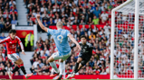 A football player in a red jersey defends against a light blue jersey player attempting to score a goal, with a goalkeeper poised to react, in front of a crowded stadium.