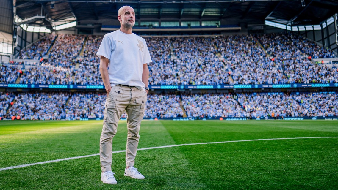 A person stands on a soccer field wearing a white Puma t-shirt and beige pants, with a large crowd in the stadium.