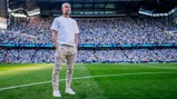 A person stands on a soccer field wearing a white Puma t-shirt and beige pants, with a large crowd in the stadium.