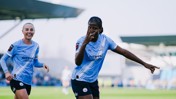 Two Manchester City Women's team players celebrate a goal on the field. One player is pointing forward. They are wearing light blue kits with the Etihad Airways logo.