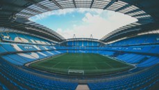 An empty football stadium with blue seats and green pitch under a partially cloudy sky.