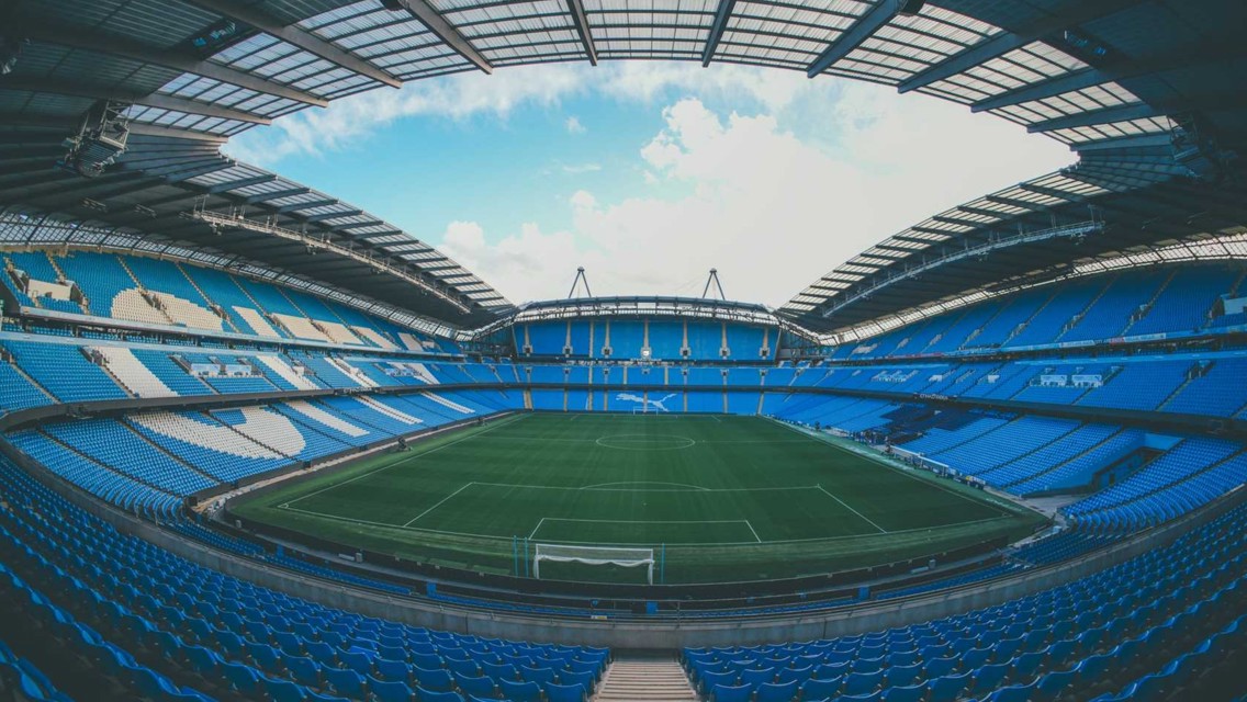 An empty football stadium with blue seats and green pitch under a partially cloudy sky.