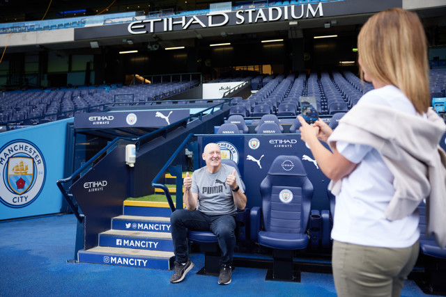 A person having their photo taken while sitting in the Manchester City dugout at Etihad Stadium.