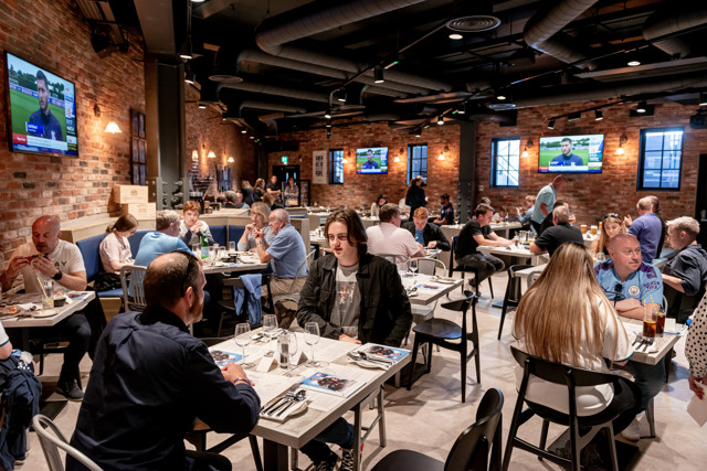 A restaurant with patrons dining at tables. Multiple TVs showing a sports broadcast with Sky Sports logo visible. Exposed brick walls and industrial decor.