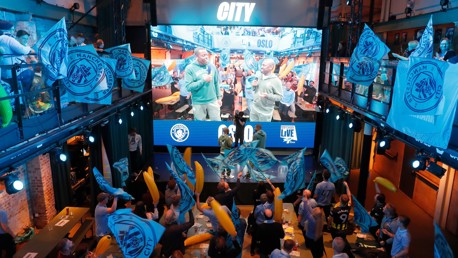 Indoor fan event featuring Manchester City flags, a large screen, and a crowd of supporters holding inflatable items.