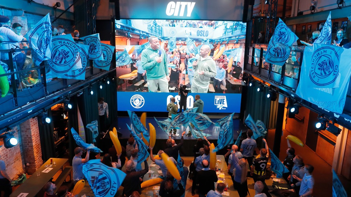 Indoor fan event featuring Manchester City flags, a large screen, and a crowd of supporters holding inflatable items.