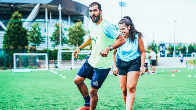 Two people on a soccer field, one wearing a Puma bib and the other a 'Citizens giving' shirt, engage in training exercises. Soccer goals and a stadium are visible in the background.