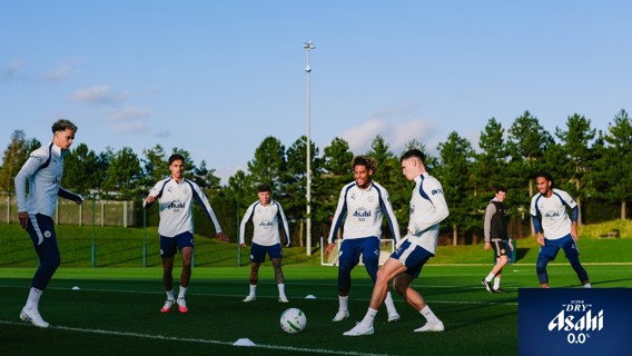 Manchester City players in training on a grassy field wearing Manchester City attire with Asahi Super Dry logo visible.