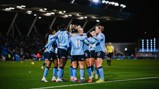 A group of athletes in light blue jerseys celebrating on a soccer field at night, with a stadium and spectators in the background.