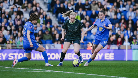 Soccer match with players in blue and black kits competing for the ball, surrounded by spectators in a stadium setting.