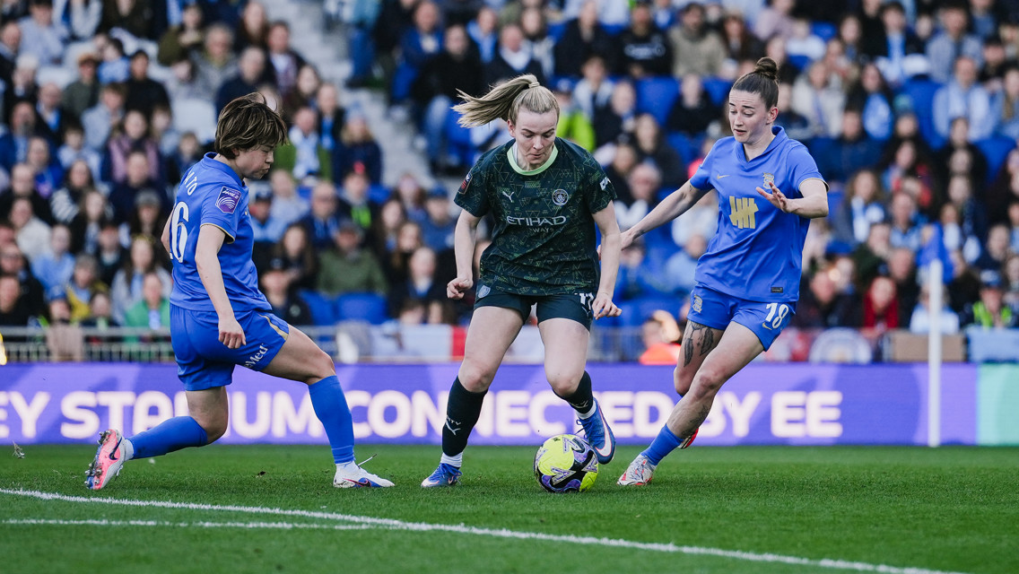Soccer match with players in blue and black kits competing for the ball, surrounded by spectators in a stadium setting.