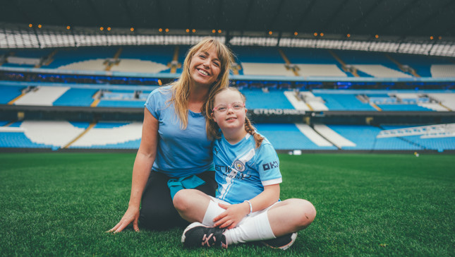 A woman and child sit on the grass at a football stadium. The child is wearing a Manchester City football kit.
