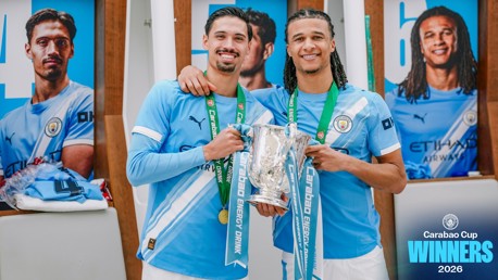 Two Manchester City players celebrate winning the Carabao Cup 2026, holding the trophy with medals in the locker room.