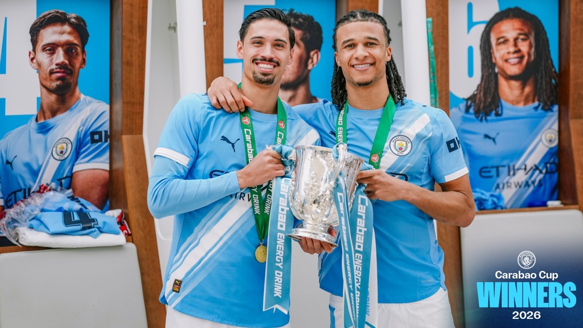 Two Manchester City players celebrate winning the Carabao Cup 2026, holding the trophy with medals in the locker room.