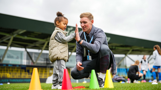 A coach kneels on a soccer field, giving a high-five to a child standing beside colorful cones. Both their faces are blurred. In the background, other children and adults are visible, likely participating in a sports event.